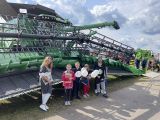 Kobieta w okularach i grupa dzieci z białymi balonami. Za nimi duży zielony kombajn