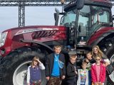 Uczniowie szkoły podstawowej i kobieta w okularach. Za nimi duży czerwony ciągnik