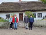 Czterech gimnazjalistów w kolorowych kurtkach. Za nimi stara drewniana chałupa z dachem pokrytym strzechą
