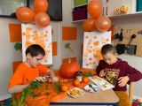 Dwóch uczniów szkoły podstawowej siedzących przy stole. Na stole pomarańczowe warzywa. Za nimi pomarańczowe balony