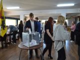 Uczeń liceum w ciemnym garniturze wrzuca kartkę do plastkiowej, przezstroczystej urny wyborczej. Obok przechodzą dwie...