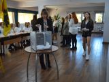 Uczennica liceum w okularach wrzuca kartkę do plastkiowej, przezstroczystej urny wyborczej. W oddali stoi grupa osób.
