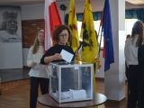 Kobieta w okularach wrzuca kartkę do plastkiowej, przezstroczystej urny wyborczej. W tle flagi: biało-czerwona, granatowa,...