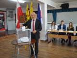 Mężczyzna w garniturze wrzuca kartkę do plastkiowej, przezstroczystej urny wyborczej. W tle flagi: biało-czerwona,...