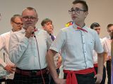 Dwóch uczniów szkoły podstawowej w białych koszulach z czerwonymi wstążkami przy kołnierzach. Jeden z nich śpiewa do...