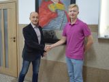 Mężczyzna w ciemnej marynarce składa gratulacje uczniowi liceum. Uczeń ma bląd włosy, ubrany jest w różową koszulkę.