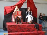 Uczenica liceum w kostiumie królowej i uczeń liceum w kostiumie króla siedzą na krzesłach przy ścianie.