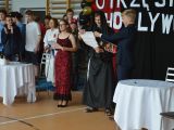 Uczeń liceum w czarnym habicie czyta z kartki. Obok stoi licealista w garniturze i licealistka w czerwonej sukience. W tle...