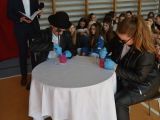 Opis alternatywny: Przy stoliku przykrytym białym obrusem siedzą dwie uczennice liceum w czarnych kurtkach. Wyciskają sok w...
