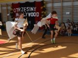 Dzewczyna i chłopak w białych pelerynach biegną środkiem sali. pod ścianą, na podłodze siędzą uczniowie liceum.