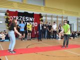Uczeń liceum z zawiązanymi oczami rzuca małą piłeczkę, naprzeciwko stoi dziewczyna z koszykiem. W tle uczniowie liceum.