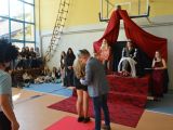 Uczenica liceum w kostiumie królowej i uczeń liceum w kostiumie króla siedzą na krzesłach. Przed nimi mężczyzna ucina ogon...