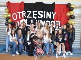 Przy ścianie grupa uczniów liceum w kolorowych ubraniach. Drugi rząd stoi, pierwszy siedzi na krzesłach. Pośród uczniów...