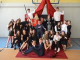 Przy ścianie grupa uczniów liceum. Drugi rząd stoi, pierwszy siedzi na krzesłach. Pośród dwoje uczniów w strju króla i...