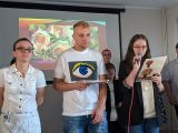 Gimnazjalista i dwie gimnazjalistki w strojach galowych. Gimnazjalistka z długimi włosami czyta do mikrofonu