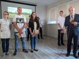 Mężczyzna w garniturze mówi do mikrofonu. Obok grupa gimnazjalistów w strojach galowych