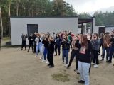 Duża grupa licealistów tańczy na plaży. W tle szary budynek i jezioro.