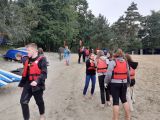 Grupa licealistów w pomarańczowych kamizelkach stoi na plaży. W tle drzewa.