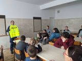 Policjantka z trzymaną przed sobą odblaskową kamizelką z napisem policja. Przed nią grupa uczniów siedzących na krzesłach