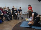 Na scenie uczennica liceum uciska klatkę piersiową manekinowi. Zdarzeniu przygląda się grupa uczniów siedzących na krzesłach.