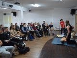 Na scenie uczennica liceum uciska klatkę piersiową manekinowi. Zdarzeniu przygląda się grupa uczniów siedzących na krzesłach.