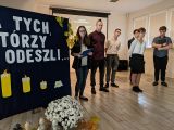 Czworo gimnazjalistów w strojach galowych. Obok granatowa tablica z napisem Dla tych, którzy odeszli