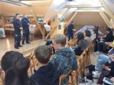 Siedząca grupa młodych ludzi otrzymuje broszurki od policjantki w granatowym mundurze.
