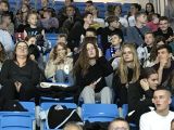 Uczniowie liceum, kobiety i mężczyźni siedzą na trybumach i obserwują mecz piłki ręcznej, który rozgrywany jest na boisku.