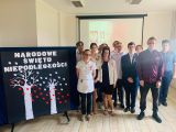 Grupa giminazjalistów w strojach galowych i biało-czerwonymi wstążkami na piersi. Obok granatowa tablica z napisem Narodowe...