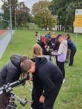 Na pierwszym planie chłopiec z dziewczyną patrzą przez teleskop. Inna grupa młodzieży stoi przy teleskopach.