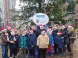 Pięć kobiet w średnim wieku i grupa uczniów w czerwonych czapkach. W tle duża choinka przystrojona oświetleniem neonowym