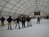 Duża grupa uczniów liceum jeździ na łyżwach po krytym lodowisku. Pięć osób trzyma się za ręce. Wszyscy są uśmiechnięci.