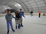 Troje uczniów liceum trzyma się za ręce i jeździ na łyżwach po krytym lodowisku. Wszyscy są uśmiechnięci.