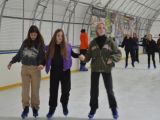 Duża grupa uczniów liceum jeździ na łyżwach po krytym lodowisku. Trzy dziewczyny trzymają się za ręce. Wszyscy są...