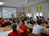 Uczniowie liceum siedzą w czterech grupach przy stolikach w sali lekcyjnej. Pośrodku stoi dziewczyna i wkazuje coś ręką.