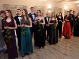 Uczennice liceum w sukniach balowych stoją w rzędzie. Za nimi stoją chłopcy. W rękakach każda dziewczyna trzyma czerwoną...