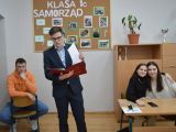 Uczeń liceum w garniturze i okularach stoi pośrodku sali. Z oku przy ławce siędzą dwie uczennice, z drugiej strony uczeń...