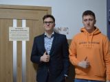 Przy drzwiach stoi obok siebie dwóch uczniów liceum. Jeden jest w garniturze i okularach, drugi w pomarańczowej bluzie.