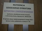Na drzwiach wisi kremowa kartka z napisami. Napisy są w kolorze czarnym i fioletowym.
