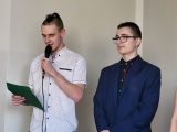 Gimnazjalista w białej koszuli w kropki czyta z kartki trzymając mikrofon. Obok chłopak w granatowym garniturze i okularach