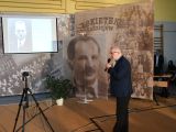 Starszy mężczyzna stoi pośrodku sali i mówi do mikrofonu. Na ścianie przed nim wiszą stare zdjęcia i ekran projekcyjny.