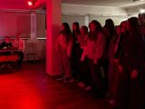 Duża grupa dziewczą z liceum stoi pośrodku sali, jedna z nich trzyma mikrofon i śpiewa. Z lewej strony przy stoliku siedzi...