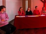 Przy stoliku przykrytym czerwonym obrusem siedzą cztery kobiety. Z lewej strony siedzi chłopak w różowej bluzie.