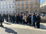 Duża grupa uczniów liceum stoi na placu. Wszyscy ubrani są w kurtki. W tle duży budynek.