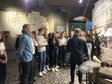 W sali muzealnej duża grupa licealistów słucha stojącego przed nimi mężczyzny w czarnej koszuli.