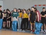 Przy ścianie w sali gimnastycznej stoi duża grupa uczniów liceum. Wśród nich trzy osoby stoją w niebieskich workach.