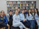 Ośmioro uczniów liceum siedzi na krzesłach. Wszyscy mają niebieskie bluzy. W tle regały z książkami.