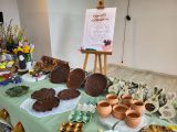 Ceramiczne kolorowe talerze, kubki i zające na długim stole przykrytym seledynowym obrusem. W tle plakat z napisem Kiermasz...