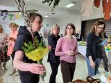 Kobieta w średnim wieku z gałązkami wierzby w dłoni. Obok trzy kobiety oglądają wielkanocne ozdoby leżące na stole
