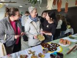 Trzy uśmiechnięte kobiety w średnim wieku. Przed nimi stół nakryty białym obrusem z glinianymi kolorowymi przedmiotami